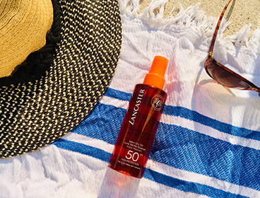 Huile solaire corps Lancaster SPF50 avec une chapeau et une serviette de plage.